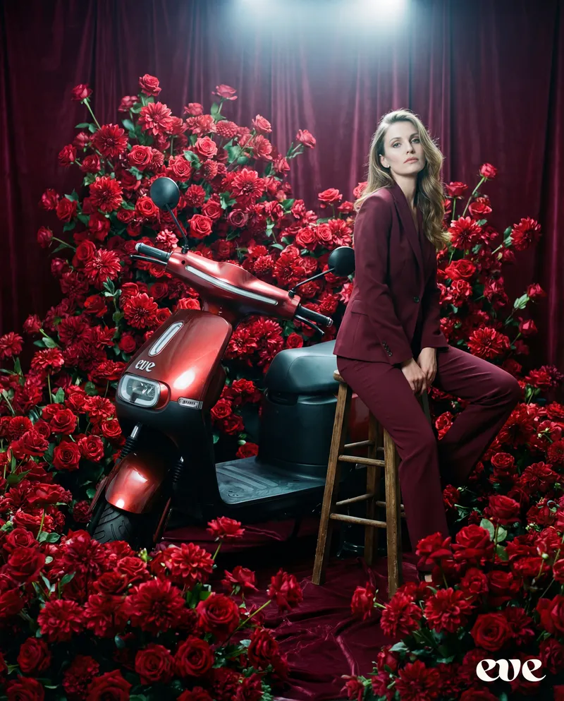 Woman beside a red EVE scooter surrounded by deep red roses — zero-emission riding in bloom